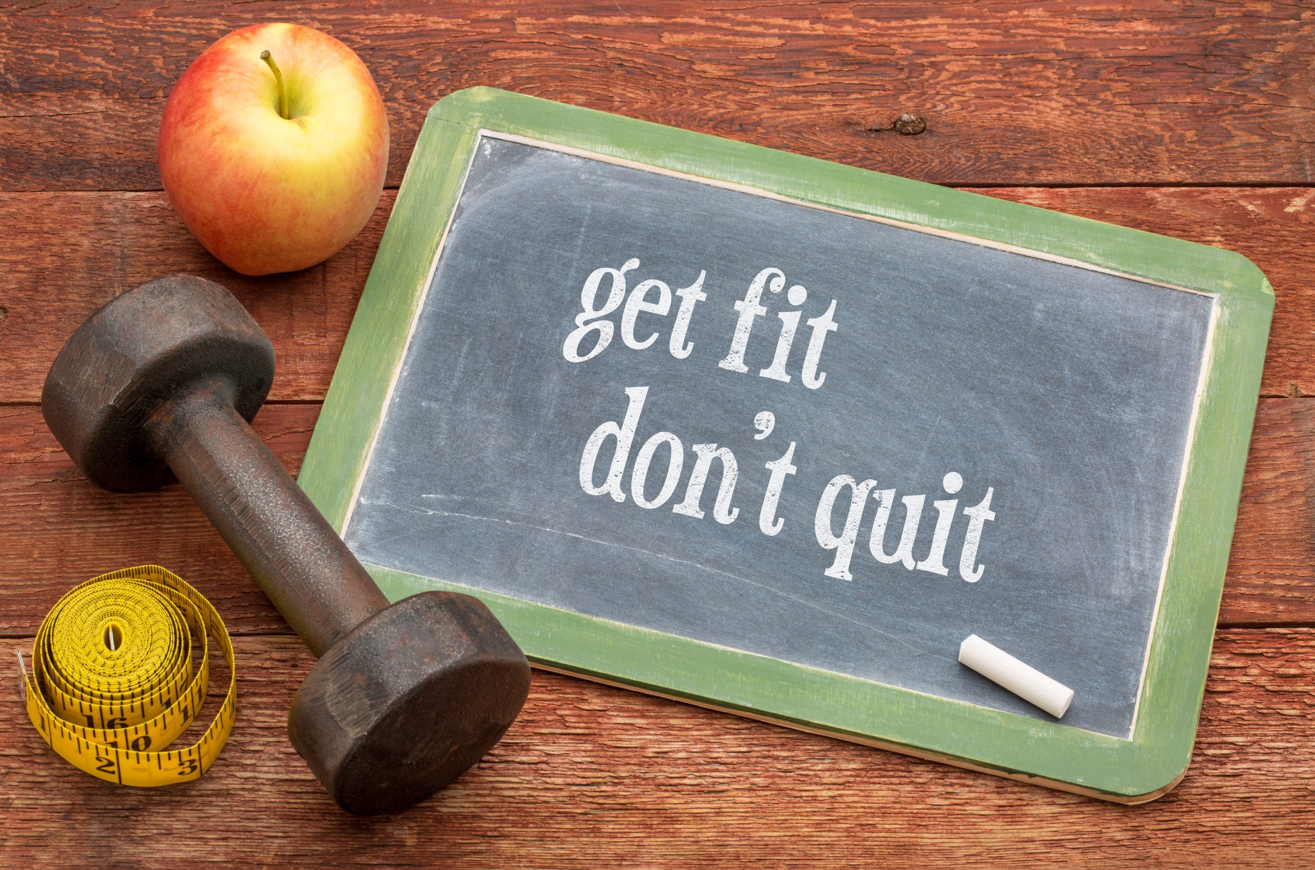 Kleine Tafel, auf der "get fit don´t quit" steht mit einer Kreide, einem Apfel, einer Hantel und einem Maßband