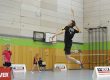 Das Bild zeigt eine Halle mit dem Fokus auf Jonas Grün, welcher einen Jump Smash macht. Im Hintergrund sind weitere Leute die Badminton spielen. Es sind Banner von Victor und Oliver zu sehen.