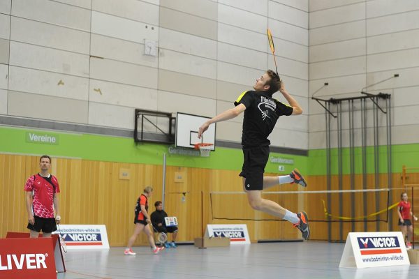Das Bild zeigt eine Halle mit dem Fokus auf Jonas Grün, welcher einen Jump Smash macht. Im Hintergrund sind weitere Leute die Badminton spielen. Es sind Banner von Victor und Oliver zu sehen.
