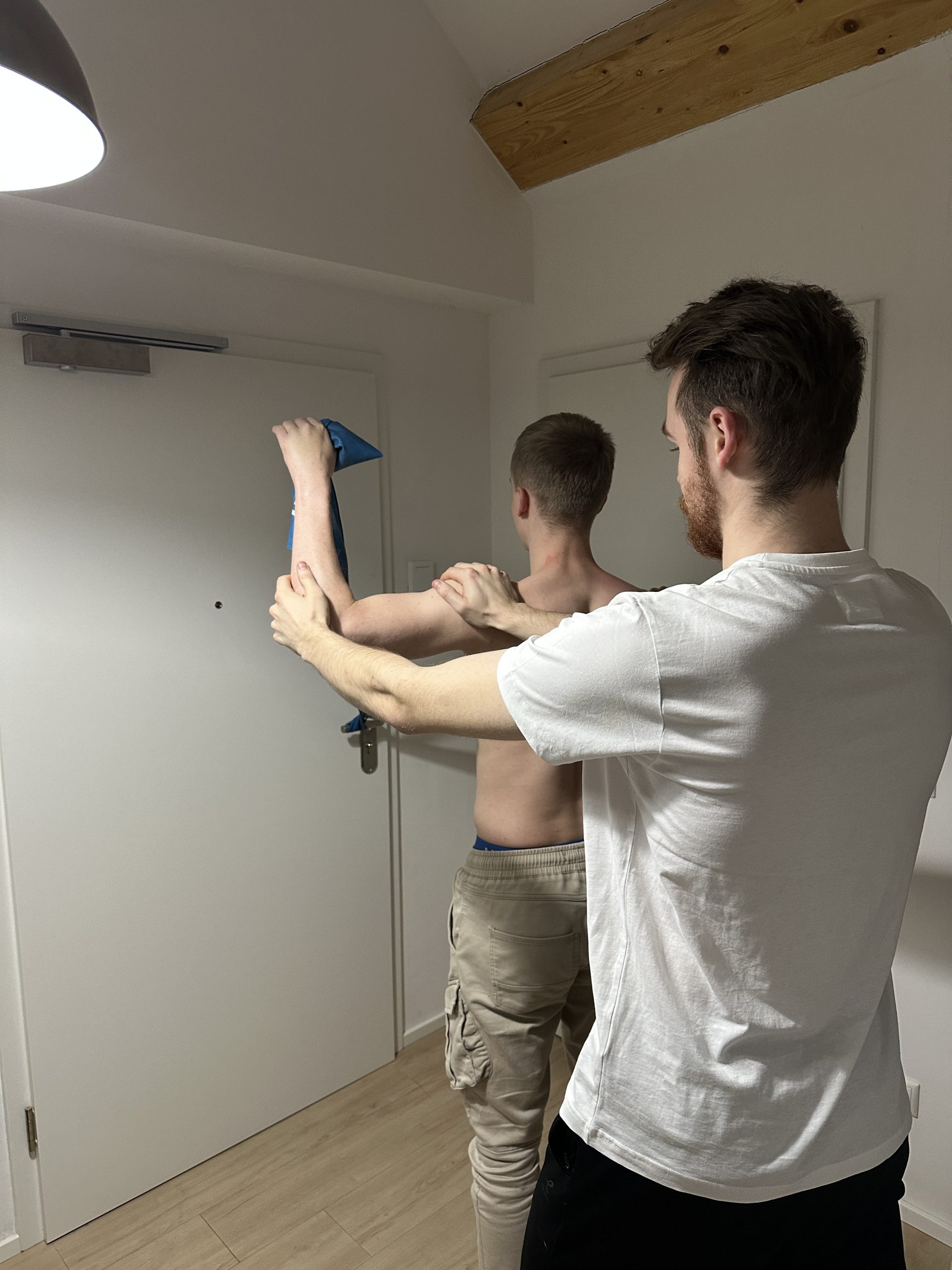 Jonas Grün, der Inhaber von Physiotherapie Grün, steht auf der rechten Seite des Bildes. Er steht in einem Raum mit weißen Wänden und im linken Bildbereich sind zwei Türen zu sehen. Jonas steht direkt vor einem Patienten und hat eine Hand auf dessen Schultern gelegt. Die andere Hand hält den Ellenbogen fest. Der Patient führt gerade eine Übung mit einem blauen Theraband aus. Dabei ist das Theraband an der Türklinke befestigt und der Patient zieht seine Arme im Rechtenwinkel nach hinten.